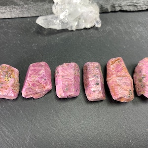 以下が含まれることがあります： 暗い灰色の表面に、自然な形をしたピンクのルビーの結晶が6つ並んでいます。結晶はすべてほぼ同じ大きさで、わずかに半透明に見えます。