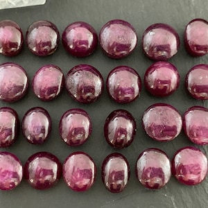 May include: A collection of twenty-three polished, cabochon-cut rubies arranged in four rows. Each gemstone features a deep, saturated magenta-red hue with a smooth, rounded surface and visible internal inclusions characteristic of natural rubies.