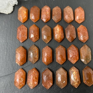May include: A collection of faceted sunstone gemstones. The stones are a warm orange-brown color with sparkling inclusions. The gemstones are cut into a hexagonal shape with a flat top and bottom. A cluster of clear quartz crystals is in the upper left corner.