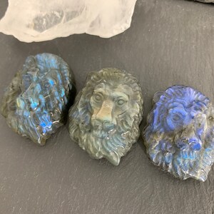 May include: Three carved labradorite lion head sculptures. The sculptures are a dark blue-grey color with iridescent flashes of blue and green.