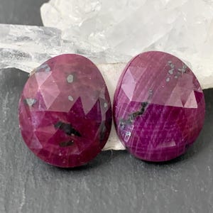 May include: Two faceted, oval-shaped ruby gemstones. The rubies are a deep, rich red with dark inclusions and a polished, reflective surface. They are set against a backdrop of a white crystal and a dark gray surface.