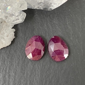 May include: Two faceted, teardrop-shaped, deep red ruby gemstones. The gems are displayed on a dark gray surface, with a cluster of white, crystalline material in the background.