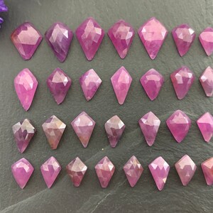 May include: A selection of pink and purple faceted gemstones, cut into diamond shapes, displayed on a dark grey surface. The gemstones vary in size and shade, highlighting their geometric facets and natural colour variations. A few purple flower petals are visible.
