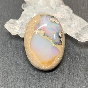 May include: Oval-shaped gemstone with a tan, speckled border. The center features a mix of colors, including white, blue, red, and green. A clear crystal formation is visible in the background. The gemstone is likely a pendant or a decorative element.