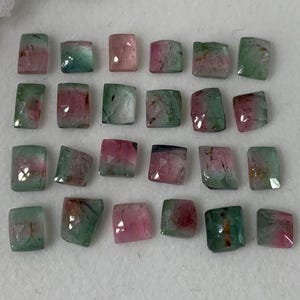 May include: A collection of rectangular watermelon tourmaline gemstones. The stones display a mix of pink and green hues, with some having a clear division of color. The gemstones are arranged on a white surface.