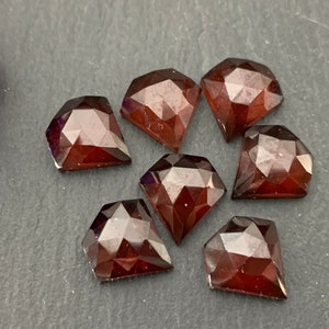 May include: Seven faceted, diamond-shaped gemstones in varying sizes. The gems are a deep, translucent red-brown colour, with multiple facets catching the light. The gemstones are arranged on a dark grey surface.