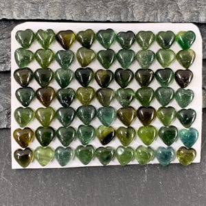 May include: A collection of heart-shaped gemstones in varying shades of green, from olive to deep forest. The polished stones are arranged on a white card, creating a visually appealing display. The gemstones are likely used for jewelry or crafts.