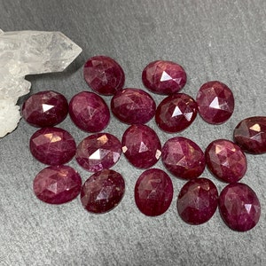 May include: A collection of 16 oval-shaped ruby gemstones with a faceted cut. The gemstones are a deep red color and have a glossy finish.
