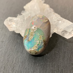 May include: An oval-shaped opal gemstone with iridescent blue, green, and red flashes. The opal is set against a backdrop of a clear quartz crystal. The stone has a rough, textured surface with a mix of colors.