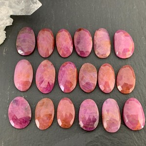 May include: A collection of oval-shaped, faceted ruby gemstones in varying shades of pink and red. The gemstones are arranged on a dark gray surface, with a clear crystal formation in the upper left corner. The gems have a polished, reflective surface.