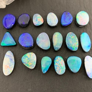 May include: A collection of polished opal gemstones in various shapes and sizes. The opals display a range of colors, including blues, greens, and whites, with iridescent flashes. The gemstones are arranged on a dark surface.