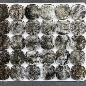 May include: A collection of thirty square-shaped rutilated quartz cabochons arranged in a grid. Each translucent stone features a unique pattern of black needle-like inclusions embedded within the clear, polished crystalline structure.
