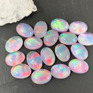 May include: A collection of oval-shaped opals with iridescent colors of pink, blue, and green. The gemstones are arranged on a dark surface, with a cluster of white crystals in the background. These opals would be suitable for jewelry making.