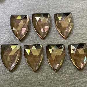 May include: Eight faceted shield-shaped gemstones with a smoky, translucent appearance. The gems have a flat back and a pointed bottom, arranged on a dark gray surface. The facets catch the light, creating a shimmering effect.