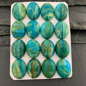 Peut inclure: Un ensemble de seize cabochons ovales polis disposés en une grille de quatre par quatre sur une carte de présentation blanche. Chaque cabochon présente un motif marbré aux teintes vives de turquoise, bleu profond et vert terreux, veiné d'or.