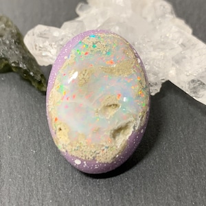 May include: Oval-shaped opal gemstone with a lavender-colored border. The opal features a white and iridescent center with flecks of color. The gemstone is surrounded by other crystals and stones.