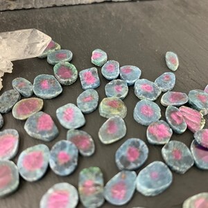 May include: A collection of small, flat, oval-shaped gemstone slices featuring a vibrant pink center surrounded by a blue-grey outer rim. A single, clear, jagged quartz crystal point is positioned at the top left of the scattered gemstone pieces.