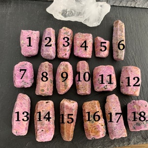 以下が含まれることがあります： 黒い表面に3列に並んだ、1から18まで番号が付けられた18個のピンクと茶色の粗いルビーの結晶。