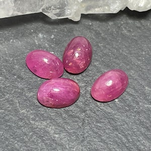 May include: Four oval-shaped, cabochon-cut rubies with a deep magenta-pink hue and a smooth, polished surface. Each gemstone displays natural internal inclusions and light reflections across its rounded top, showcasing a vibrant, saturated color.