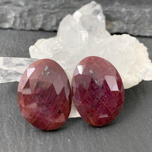 May include: Two oval-shaped, faceted ruby gemstones with a deep red color and dark inclusions. The stones have a polished, reflective surface with multiple geometric facets that catch the light, showcasing their natural texture and deep crimson hue.
