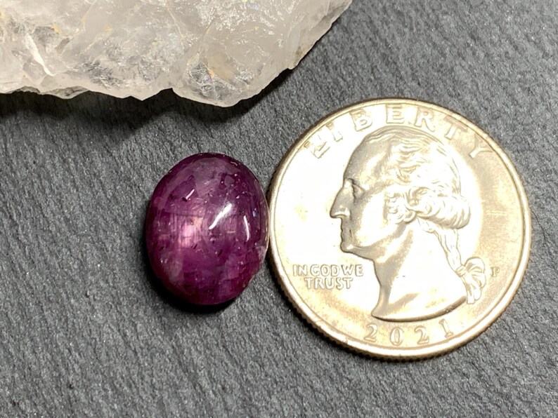 Op de afbeelding: Een gepolijste, ovaalvormige, diep magenta edelsteen naast een Amerikaanse kwart dollar munt. De edelsteen heeft een glad, reflecterend oppervlak met interne insluitsels. De munt toont het profiel van George Washington en het jaar 2021.