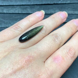 以下が含まれることがあります： 黒と緑の色合いの、涙の形をした金色の光沢のある黒曜石のカボションです。