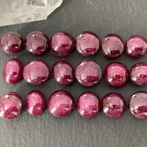May include: A collection of 18 round, cabochon cut, ruby gemstones. The rubies are a deep red color and have a smooth, polished finish.