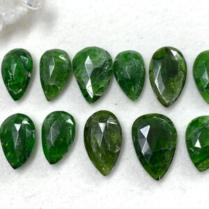 May include: A collection of eleven faceted, teardrop-shaped green gemstones. The gems vary slightly in shade, from lighter to darker greens, and are arranged on a white surface. The facets create a sparkling effect, highlighting the gems' clarity.