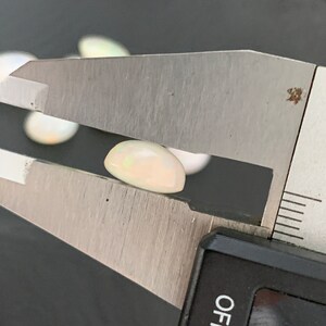 May include: A white oval-shaped opal gemstone being measured with a caliper. The caliper reads 0 to 12.