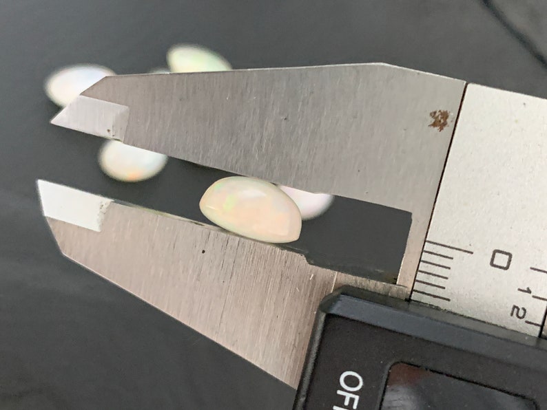 May include: A white oval-shaped opal gemstone being measured with a caliper. The caliper reads 0 to 12.