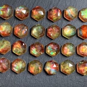 May include: A collection of twenty-four hexagonal faceted gemstones arranged in four rows of six. Each stone displays a translucent, iridescent play of color with shifting hues of orange, yellow, green, and red across the polished surfaces.