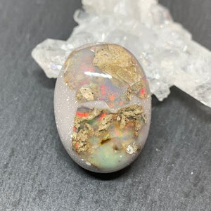 May include: Oval-shaped gemstone with a mix of colors, including red, gold, and green, set in a light gray base. The stone has a textured appearance with a clear, crystalline formation in the background. This gemstone is suitable for jewelry making.