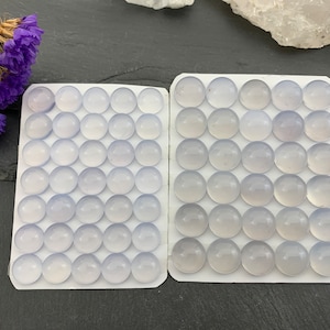 Op de afbeelding: Twee witte kaarten gevuld met ronde, lichtblauwe edelstenen. De edelstenen zijn gerangschikt in een rasterpatroon. Paarse gedroogde bloemen bevinden zich in de linkerbovenhoek. De achtergrond is een donkergrijze ondergrond.