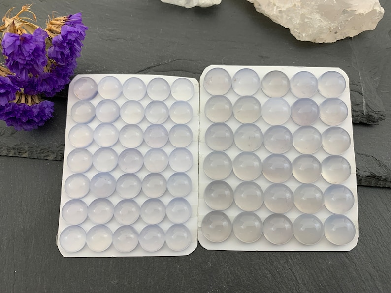 Op de afbeelding: Twee witte kaarten gevuld met ronde, lichtblauwe edelstenen. De edelstenen zijn gerangschikt in een rasterpatroon. Paarse gedroogde bloemen bevinden zich in de linkerbovenhoek. De achtergrond is een donkergrijze ondergrond.