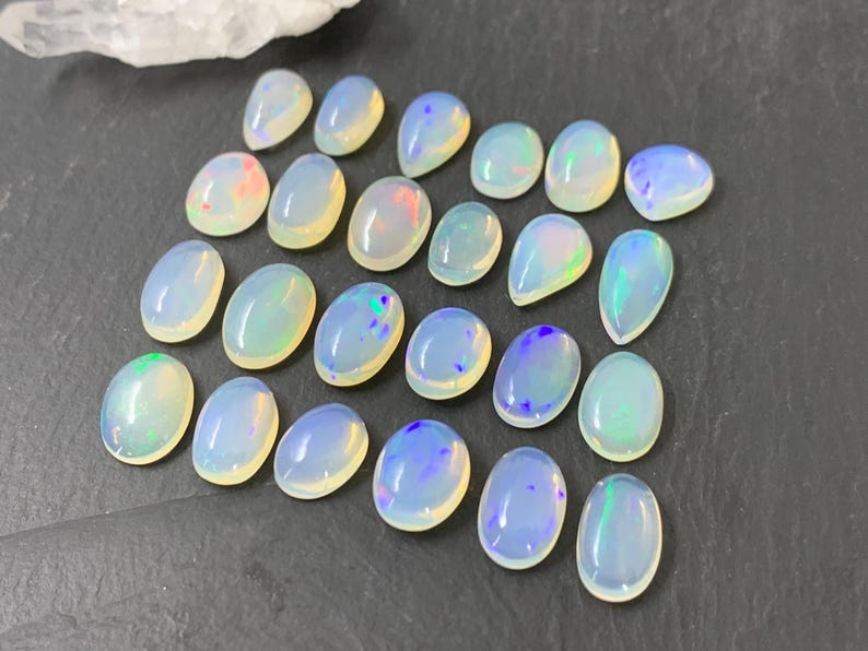 May include: A collection of 20 oval-shaped, iridescent opal gemstones. The opals are a milky white color with flashes of blue, green, and pink.