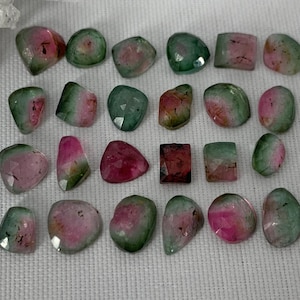 May include: A collection of 20 green, pink, and clear gemstones. The gemstones are various shapes and sizes, and have a natural, rough cut.