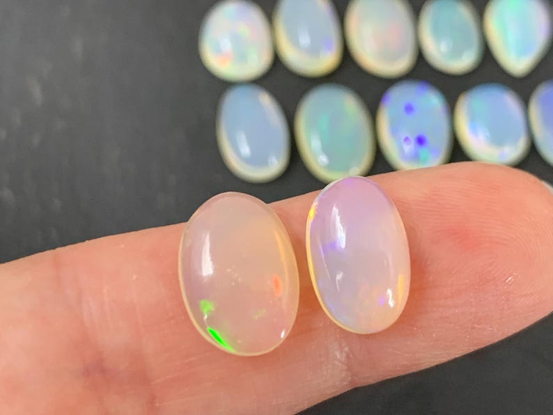 May include: Two oval-shaped, translucent, white opal cabochons with iridescent rainbow flashes. The opals are approximately 10mm in length and 7mm in width.