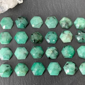 Puede incluir: Veintiuna piedras preciosas de esmeralda facetadas en forma hexagonal. Las gemas son de varios tonos de verde, algunas con inclusiones más oscuras. Están dispuestas sobre una superficie gris oscuro, con un trozo de cuarzo transparente en la esquina superior izquierda.