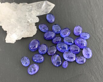 Op de afbeelding: Ovale, diepblauwe edelstenen liggen verspreid op een donkere ondergrond. Een groot, helder bergkristal bevindt zich linksboven. De edelstenen variëren in grootte en hebben een gepolijst, reflecterend oppervlak. De stenen zijn ongeveer 1-2 cm lang.