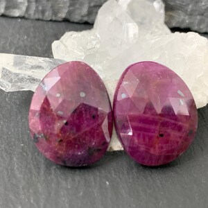 May include: Two faceted, oval-shaped ruby gemstones with a deep red hue. The gems are displayed against a backdrop of white crystal and a dark gray surface. The stones have a polished, reflective surface.