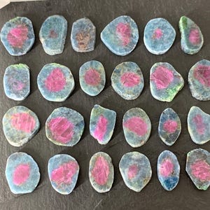 May include: Twenty-two polished gemstones with a pink center and a blue-green outer layer. The stones vary in shape and size, with a rough, natural texture. The gemstones are arranged on a dark surface.
