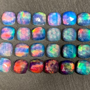 May include: Twenty-four faceted, square-cut opals with a range of colors including blue, purple, red, and green. The opals are arranged in a grid pattern on a dark gray surface. Each stone has a unique play of color.