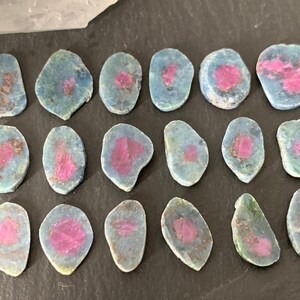 May include: A collection of 18 irregularly shaped, flat, polished stones with a blue-green base and pink accents. The stones are arranged in rows on a dark surface.