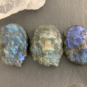 May include: Three carved labradorite lion head sculptures. The sculptures are a dark blue-grey color with iridescent flashes of blue and green.
