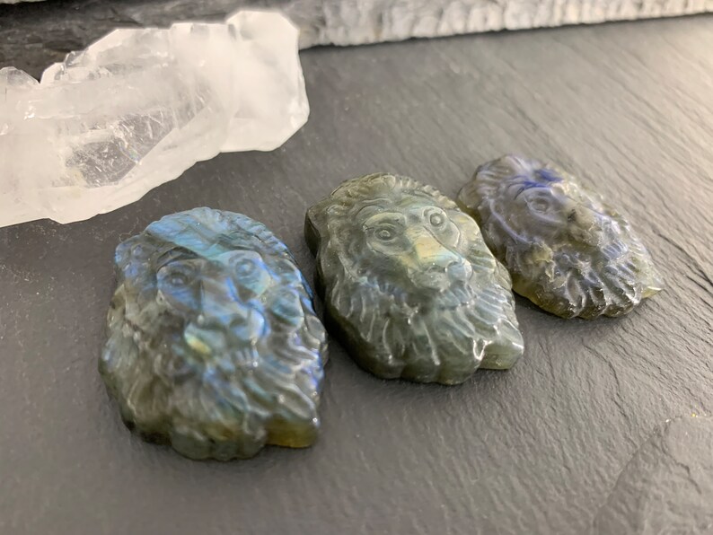 May include: Three carved labradorite lion head pendants. The pendants are a dark gray color with iridescent blue and green flashes.