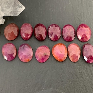 May include: An assortment of oval-shaped, faceted ruby gemstones in varying shades of red and pink. The gemstones are arranged in two rows on a dark surface, with a clear quartz crystal in the upper left corner.
