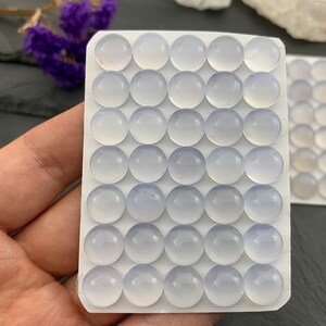 Op de afbeelding: Close-up van twee witte kaarten gevuld met ronde, lichtblauwe edelstenen. De edelstenen zijn glad en doorschijnend. De kaarten worden in een hand gehouden, met paarse bloemen op de achtergrond. Waarschijnlijk cabochons.