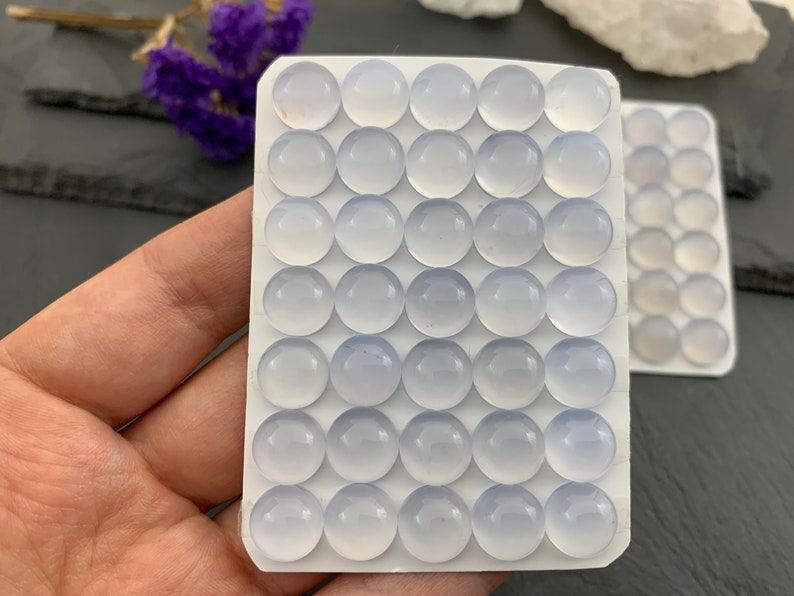 Op de afbeelding: Close-up van twee witte kaarten gevuld met ronde, lichtblauwe edelstenen. De edelstenen zijn glad en doorschijnend. De kaarten worden in een hand gehouden, met paarse bloemen op de achtergrond. Waarschijnlijk cabochons.