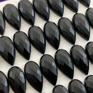 May include: A collection of black faceted teardrop-shaped gemstones. The stones are arranged in rows on a white surface.