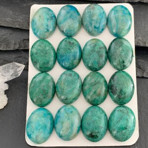 Peut inclure: Seize cabochons de forme ovale disposés en une grille de quatre par quatre sur une carte rectangulaire blanche. Chaque cabochon présente une surface polie avec un motif marbré aux teintes bleu sarcelle, vert forêt et turquoise vif.